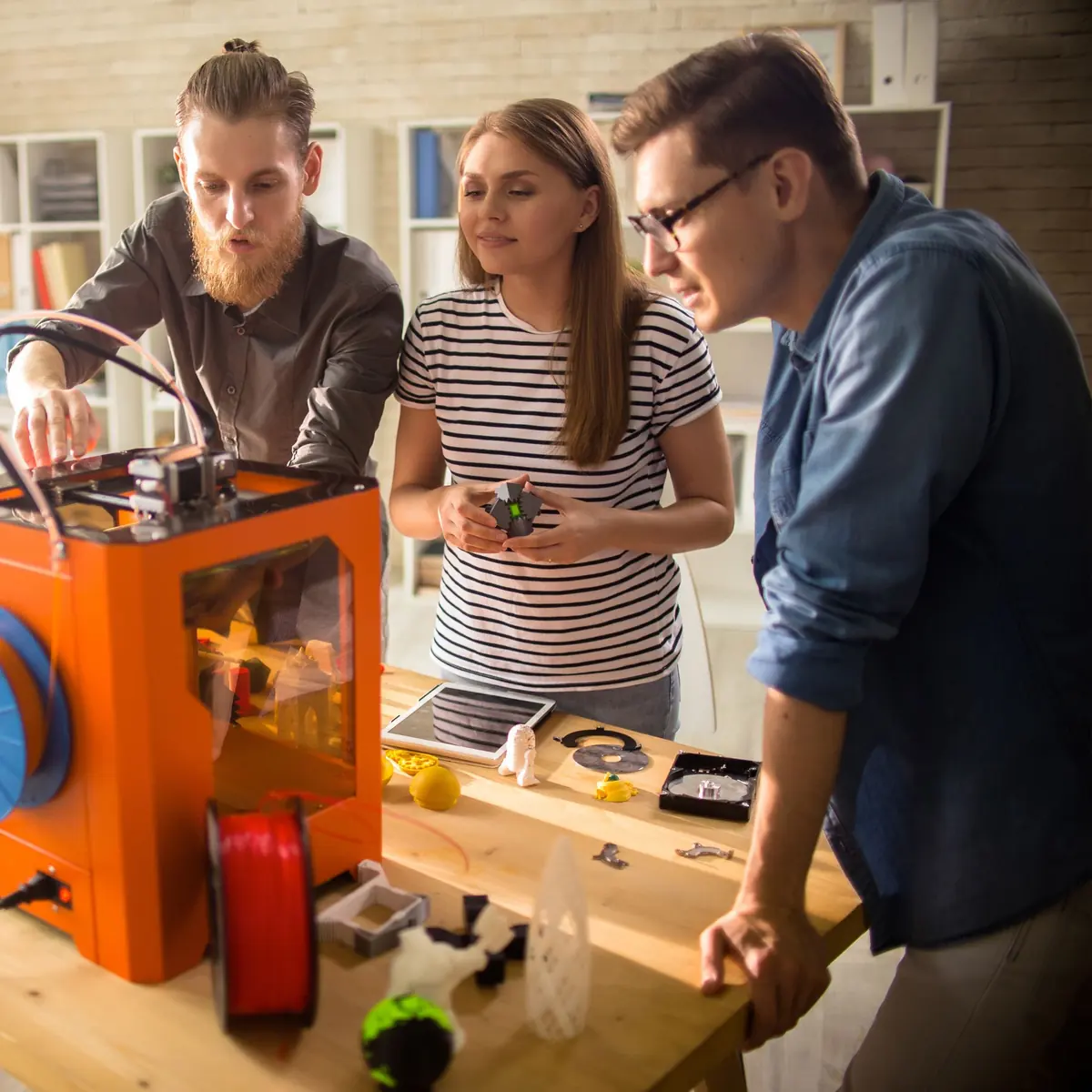 Three people watching a desktop 3D printer run beside a workbench of freshly printed parts and filament spools